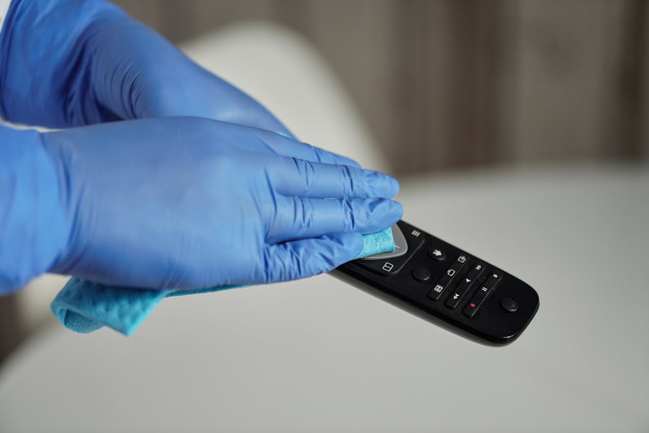 Hands in gloves sanitizing a remote control for hygiene purposes, emphasizing cleanliness.
